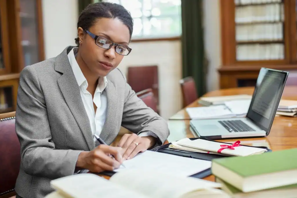 Une professionnelle RH étudie des documents réglementaires dans un bureau. Entourée de dossiers, d’ouvrages de référence et d’un ordinateur portable, elle prend des notes pour analyser les évolutions juridiques applicables à son organisation.