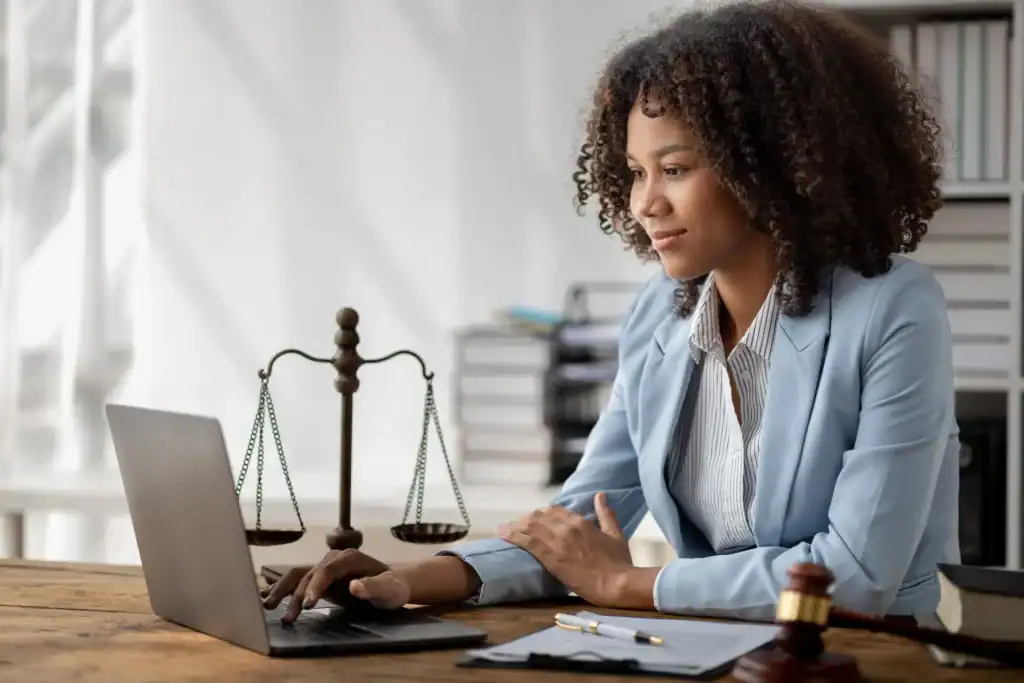 Une experte juridique consulte des informations réglementaires sur ordinateur dans un environnement de travail dédié au droit. Une balance de justice et des documents administratifs illustrent les activités de veille et d’analyse juridique.